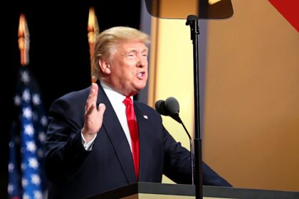 políticas de Trump reduzem crescimento da população migrante, e a imagem mostra Donald Trump discursando em um púlpito, de terno escuro e gravata vermelha, com a mão levantada e bandeiras dos Estados Unidos ao fundo