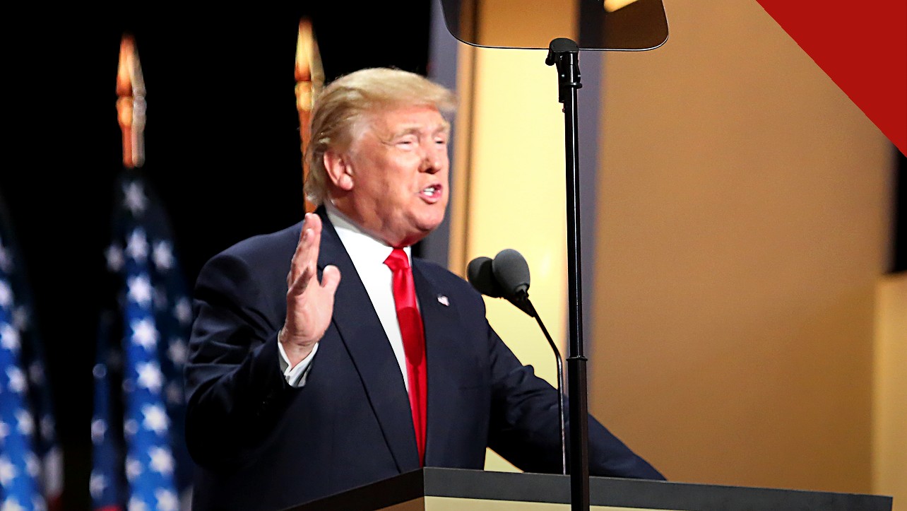 políticas de Trump reduzem crescimento da população migrante, e a imagem mostra Donald Trump discursando em um púlpito, de terno escuro e gravata vermelha, com a mão levantada e bandeiras dos Estados Unidos ao fundo
