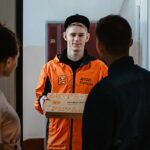 entregador arrecada mais de 500 mil dólares em vaquinha, e a imagem mostra um entregador de pizza de jaqueta laranja entregando uma caixa de pizza a um casal na porta de um apartamento, em foto de arquivo