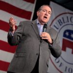 Huckabee e Carlson divergem sobre ataque de colonos a americano, e a imagem mostra Mike Huckabee discursando com um microfone na mão, de paletó cinza, diante de uma bandeira dos Estados Unidos e de um painel do Partido Republicano de Iowa
