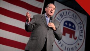 Huckabee e Carlson divergem sobre ataque de colonos a americano, e a imagem mostra Mike Huckabee discursando com um microfone na mão, de paletó cinza, diante de uma bandeira dos Estados Unidos e de um painel do Partido Republicano de Iowa