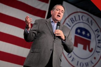 Huckabee e Carlson divergem sobre ataque de colonos a americano, e a imagem mostra Mike Huckabee discursando com um microfone na mão, de paletó cinza, diante de uma bandeira dos Estados Unidos e de um painel do Partido Republicano de Iowa