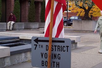 votação antecipada nos Estados Unidos bate recorde: placa que indica o local de votação do sétimo distrito nos Estados Unidos, ao lado de uma bandeira americana, com eleitores ao fundo