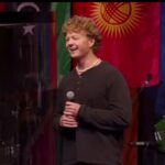 Missionário Jonathan sorri ao compartilhar o testemunho durante o mês de missões da Calvary Christian Church, com bandeiras das nações ao fundo