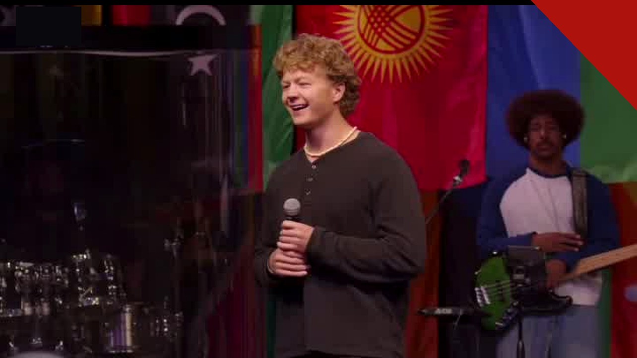 Missionário Jonathan sorri ao compartilhar o testemunho durante o mês de missões da Calvary Christian Church, com bandeiras das nações ao fundo