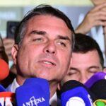 Flávio Bolsonaro, de terno escuro, fala a jornalistas cercado de microfones de emissoras em uma entrevista no Senado.