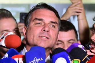 Flávio Bolsonaro, de terno escuro, fala a jornalistas cercado de microfones de emissoras em uma entrevista no Senado.