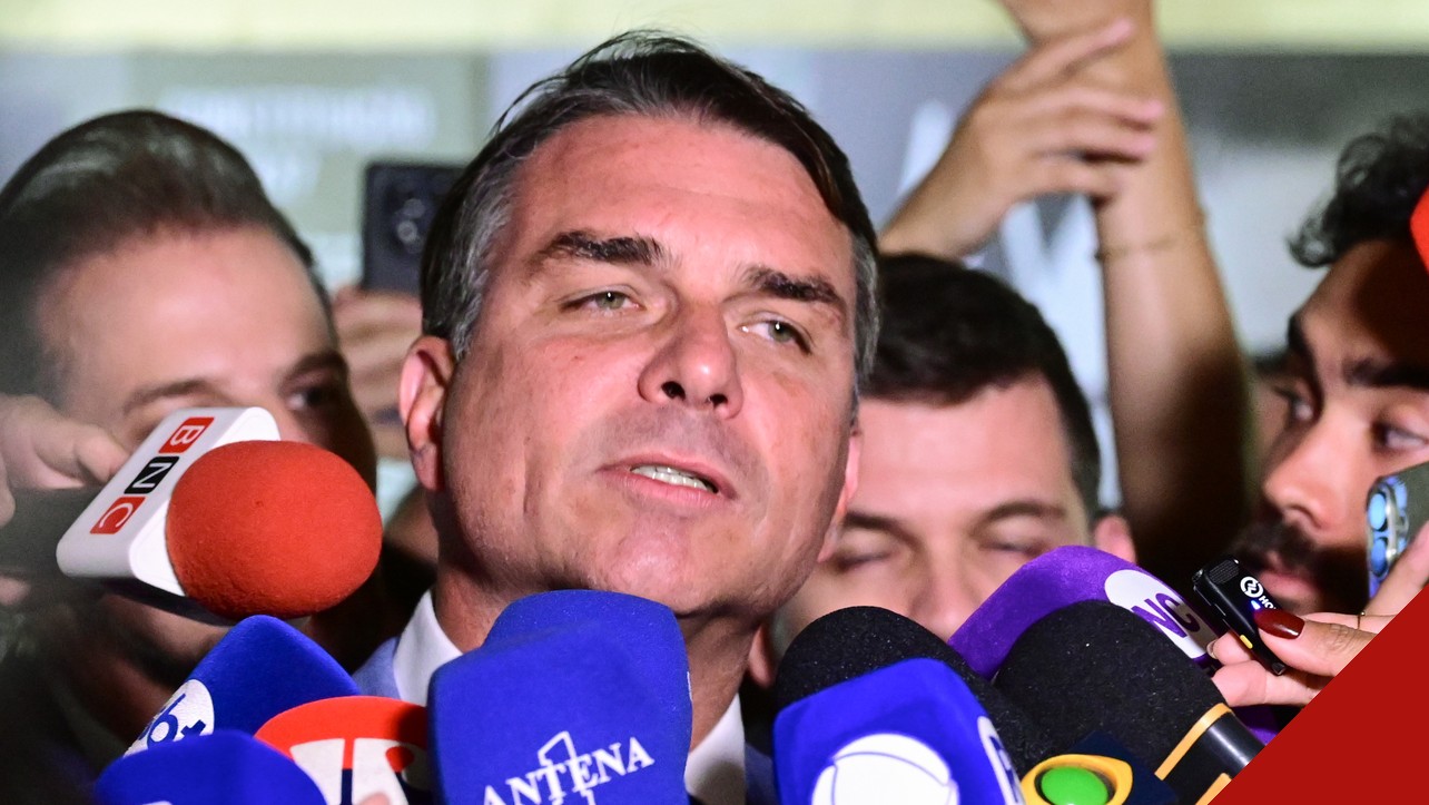 Flávio Bolsonaro, de terno escuro, fala a jornalistas cercado de microfones de emissoras em uma entrevista no Senado.
