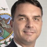 Flávio Bolsonaro, de terno escuro e gravata clara, em retrato oficial do Senado, com a bandeira do Rio de Janeiro ao fundo