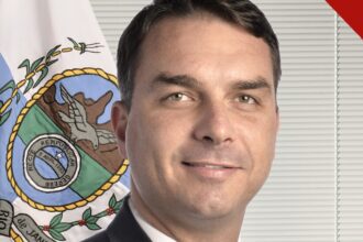 Flávio Bolsonaro, de terno escuro e gravata clara, em retrato oficial do Senado, com a bandeira do Rio de Janeiro ao fundo