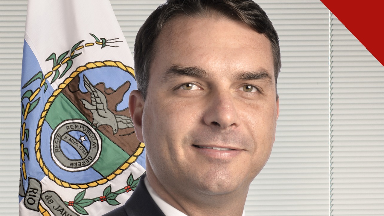 Flávio Bolsonaro, de terno escuro e gravata clara, em retrato oficial do Senado, com a bandeira do Rio de Janeiro ao fundo