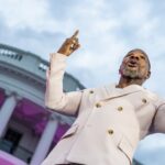 Kirk Franklin, de terno claro, aponta para o alto enquanto canta diante da Casa Branca iluminada em tons de rosa.
