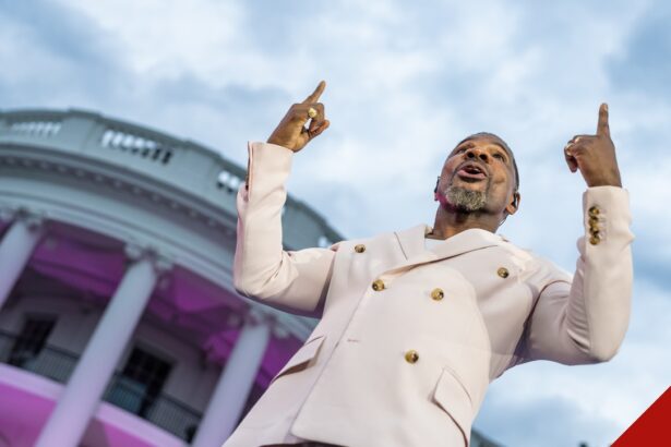 Kirk Franklin, de terno claro, aponta para o alto enquanto canta diante da Casa Branca iluminada em tons de rosa.