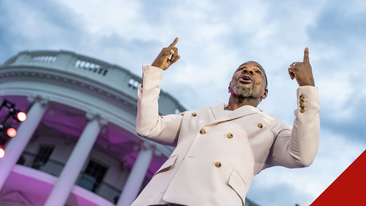 Kirk Franklin, de terno claro, aponta para o alto enquanto canta diante da Casa Branca iluminada em tons de rosa.