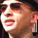 MC Guimê, de chapéu e óculos escuros, com as tatuagens do rosto e do pescoço à mostra, em retrato de 2015