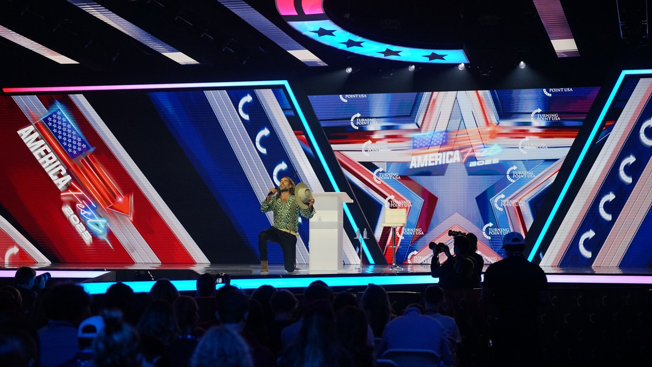 Russell Brand falando ao público no palco do AmericaFest 2025, nos Estados Unidos