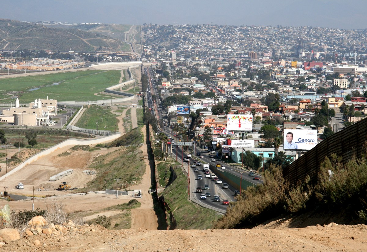 políticas de Trump reduzem crescimento da população migrante, e a imagem mostra a fronteira entre os Estados Unidos e o México, com cercas e torres de vigilância em uma área desértica, em referência ao bloqueio do fluxo fronteiriço