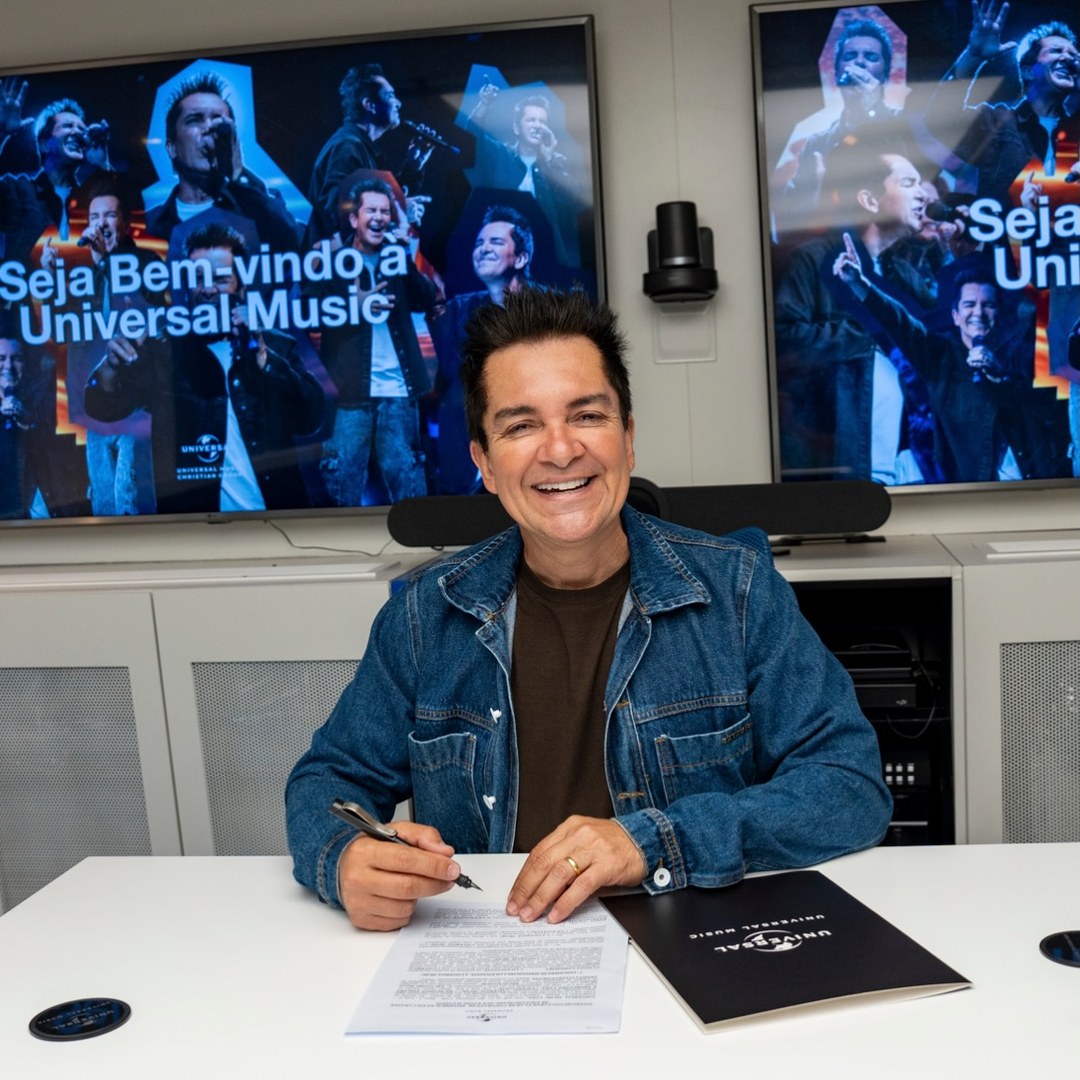 Regis Danese sentado à mesa com a caneta na mão no momento da assinatura do contrato com a Universal Music, sorrindo para a câmera