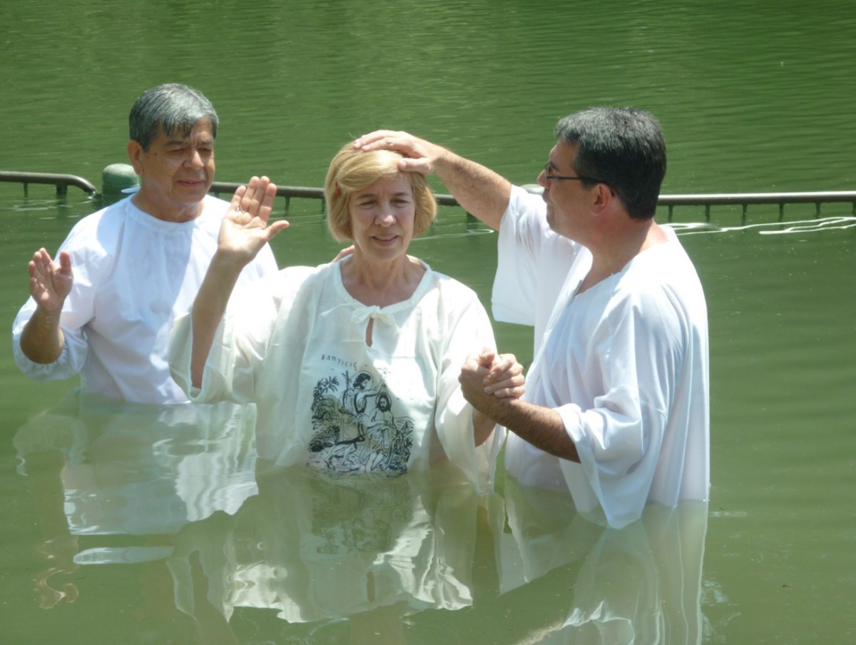Batismo nas águas realizado ao ar livre, com pastores e uma mulher de túnica branca dentro do rio, em imagem ilustrativa de batismo cristão